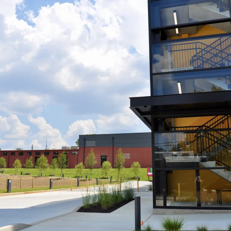 Modern glass building with visible staircases beside a green lawn and partly cloudy blue sky.