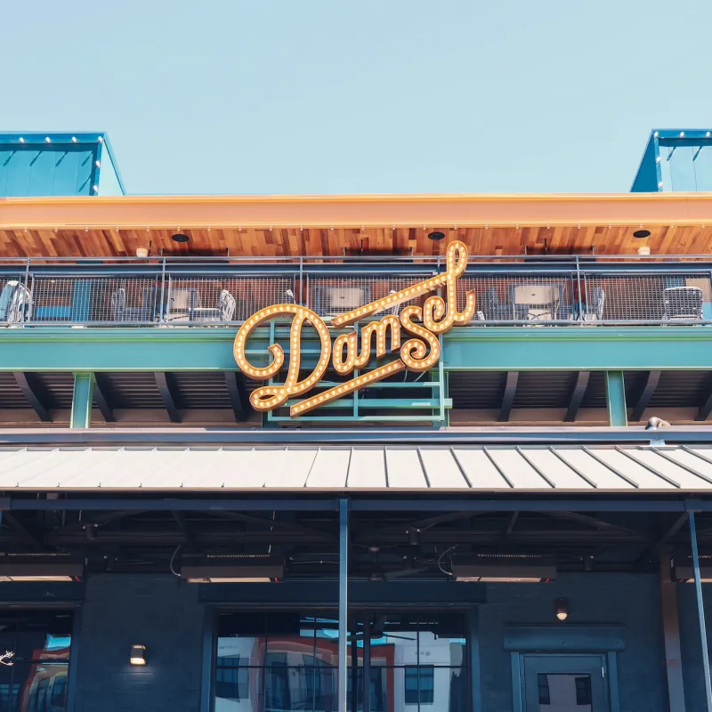 Outdoor view of Damsel restaurant with illuminated sign and rooftop seating under a clear blue sky.