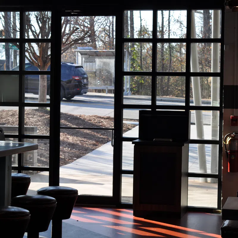 Interior view of a cafe with stools and tables near large glass windows showing an outdoor sidewalk and passing car.