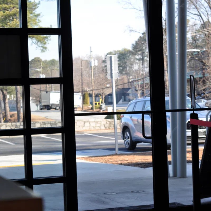 View through glass doors showing a parking lot, a parked silver SUV, and trees on a sunny day.