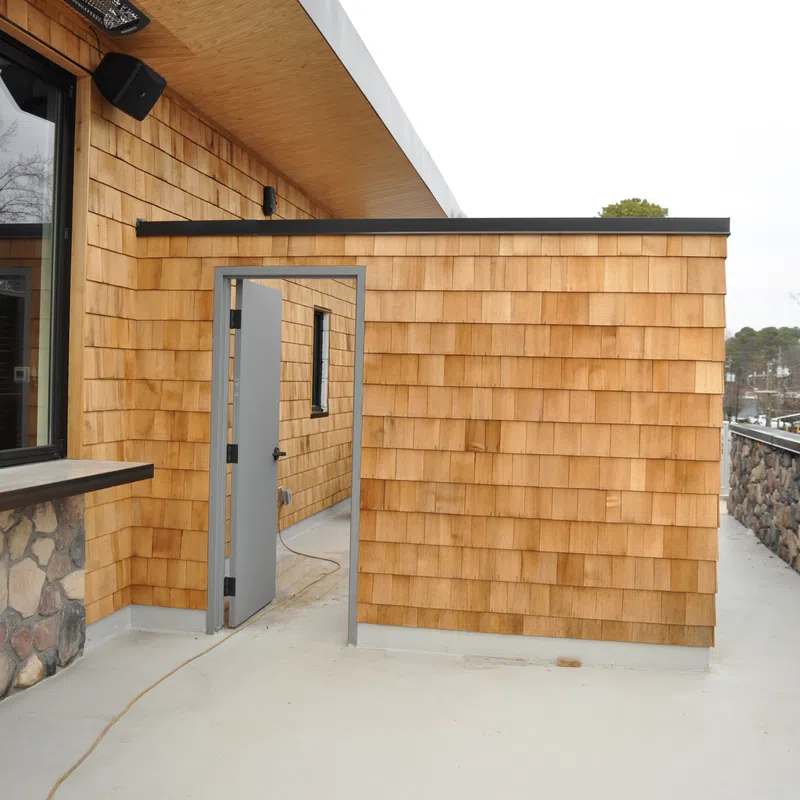 Outdoor patio area with wooden shingle walls, stone accents, glass windows, and a metal door leading inside.