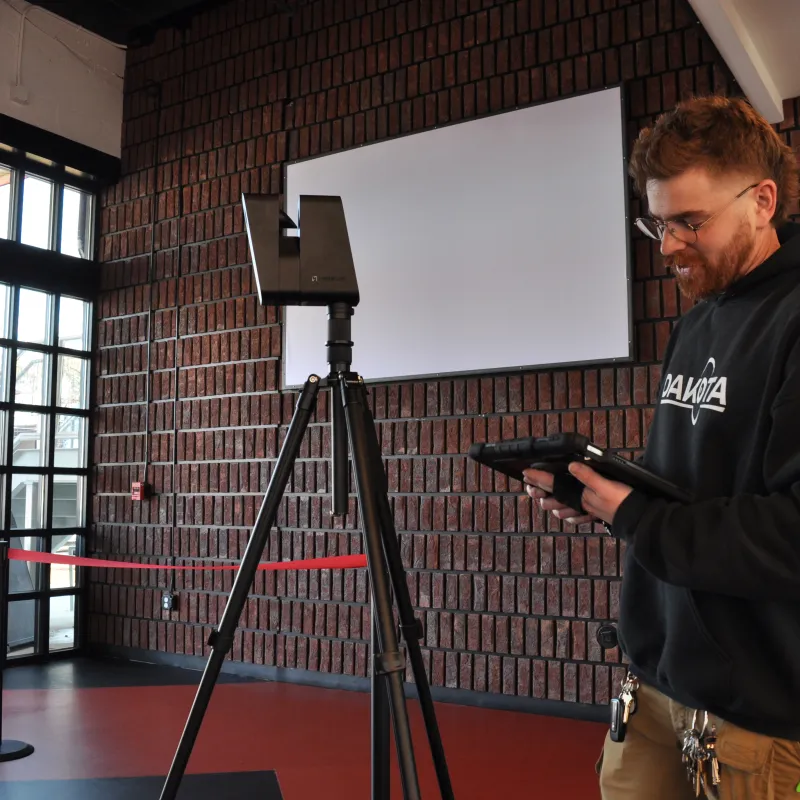 Man operating scanning equipment on tripod inside a brick-walled room with large windows and exit sign.