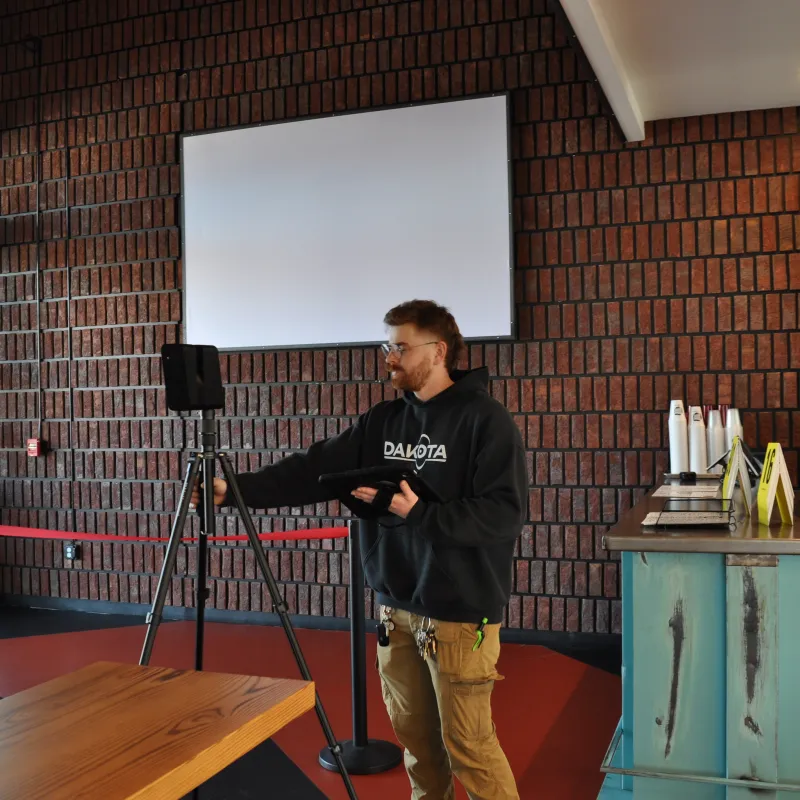Man in black hoodie setting up a speaker on a tripod inside a modern brick-walled room with large windows.