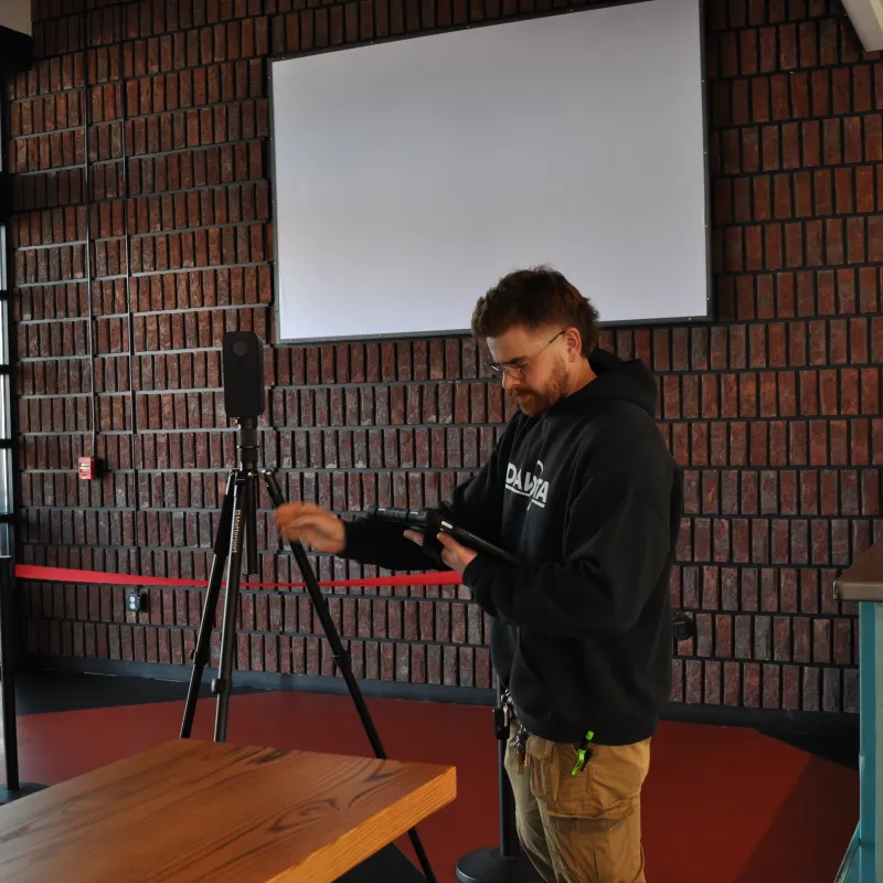 Man adjusting a microphone on a tripod in an industrial-style room with brick walls and large windows.