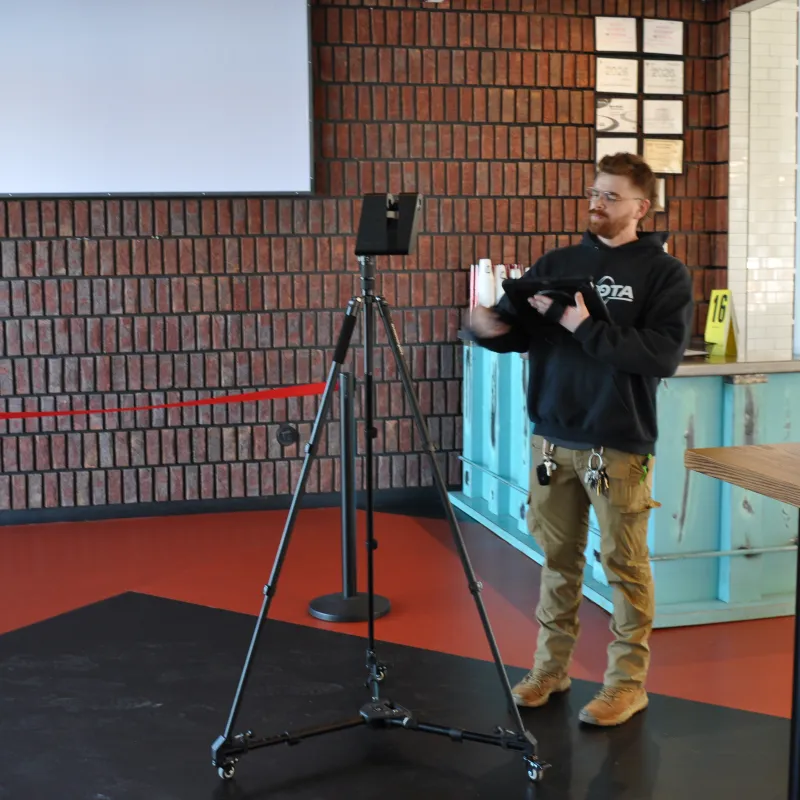 Man in casual clothing using a tablet and standing near a tripod with a device in an indoor room with brick walls.