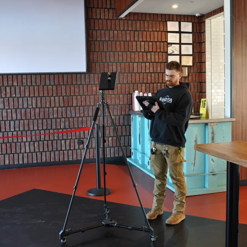 Man using a tablet to operate a tripod-mounted device in a modern indoor space with brick walls and wooden furniture.
