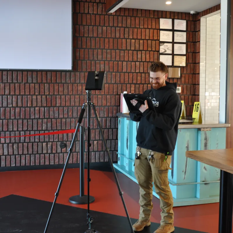 Man operating a camera setup on a tripod in a modern indoor space with brick walls and wooden furniture.
