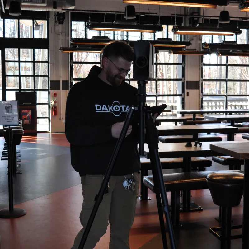 Man in black hoodie setting up camera equipment on tripod inside modern empty restaurant with large windows.