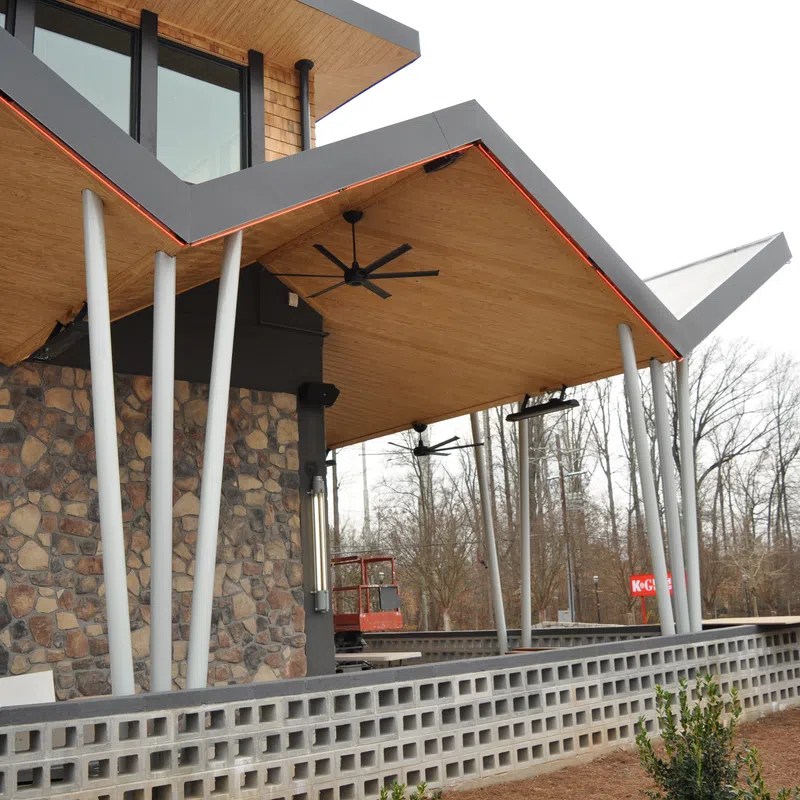 Modern building exterior with angled wooden roof, white pillars, stone wall, and outdoor ceiling fans overlooking a garden.