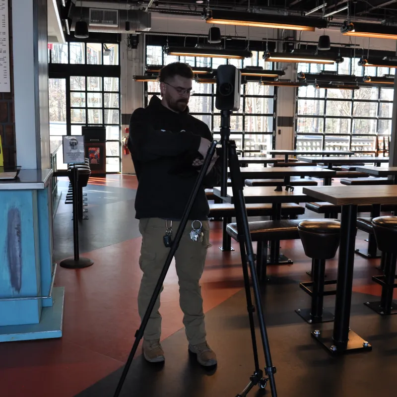 Man adjusting camera on tripod inside empty modern restaurant with wooden tables and stools.