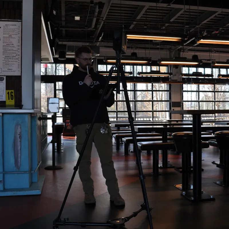 Man using camera on tripod inside a dimly lit, industrial-style cafe with large windows and empty tables.
