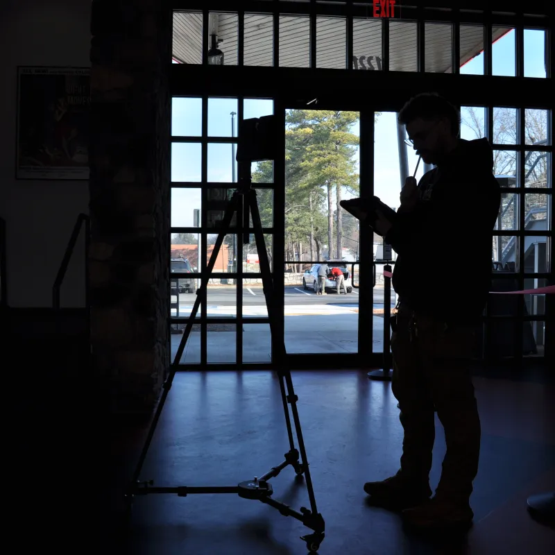 Silhouette of a person standing inside a building near an easel and glass doors showing an outdoor parking area.