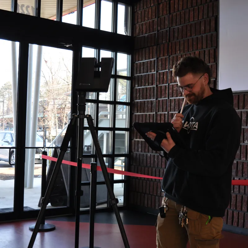 Man in casual clothes using a tablet and stylus near a tripod in a room with large windows and brick walls.