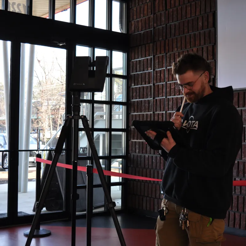 Man in a black hoodie using a tablet and stylus inside a modern building near a tripod and large windows.