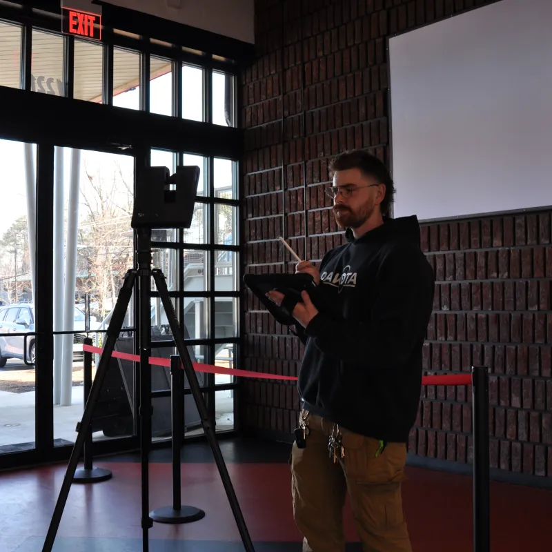 Man using a tablet near a tripod-mounted camera inside a room with large windows and brick walls.