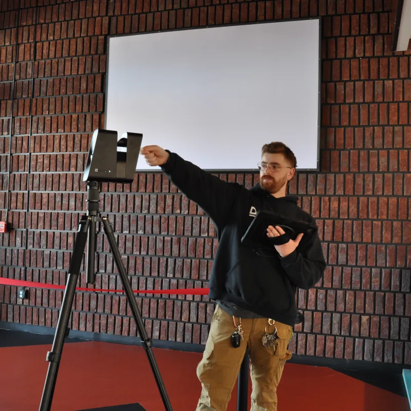 Man adjusting a large camera on a tripod inside a modern brick-walled room with large windows and red flooring.