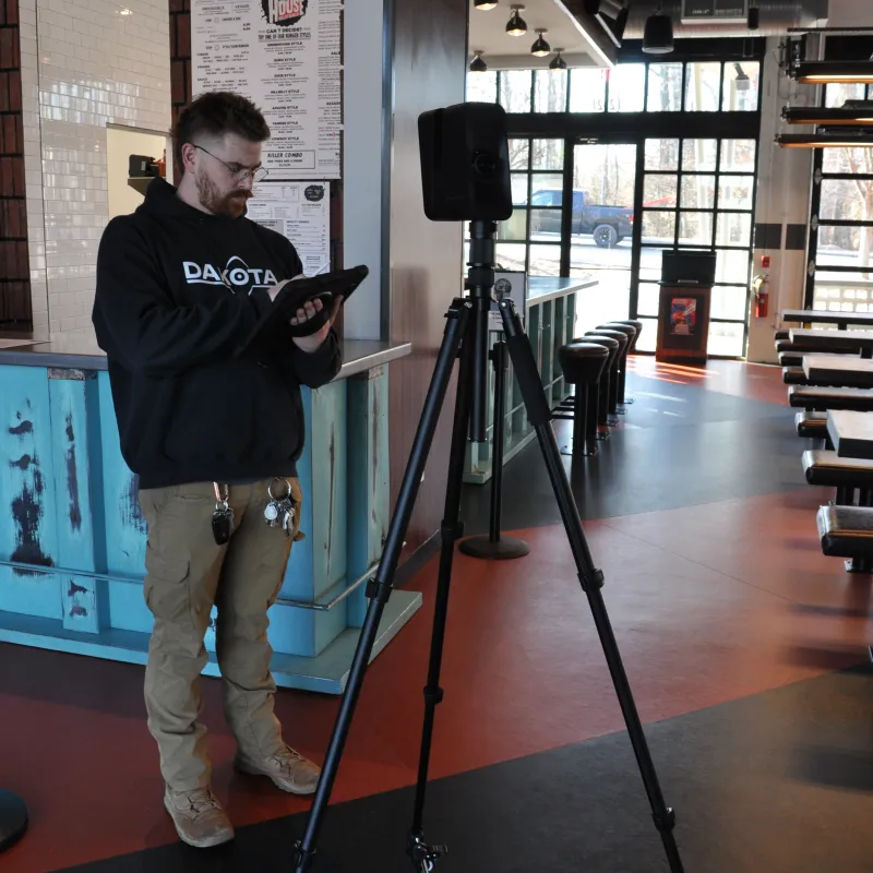 Man in a black hoodie operating a tablet near a speaker on a tripod in a modern cafe with blue counter and wooden tables.