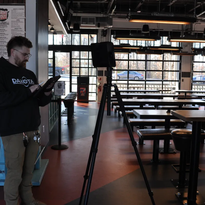Man in black Dakota hoodie using tablet in a modern empty cafe with large windows and wooden tables.
