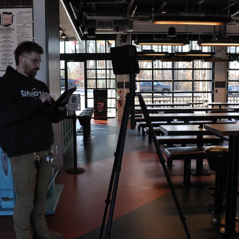 Man in casual clothing using a tablet inside a modern empty cafe with large windows and wooden tables.