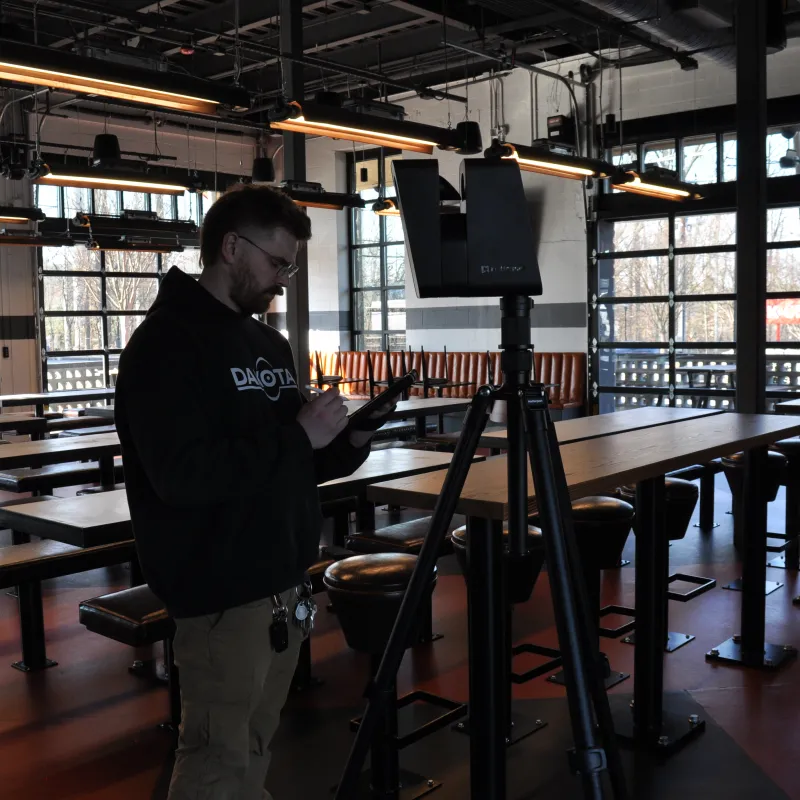 Man operating a device on a tripod in a spacious industrial-style room with large windows and wooden tables.