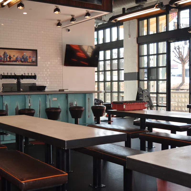 Modern indoor bar with long tables, leather benches, bar stools, large windows, and industrial lighting fixtures.