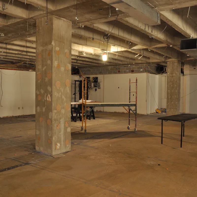 Empty unfinished basement with exposed concrete pillars, ceiling beams, and construction equipment scattered around.