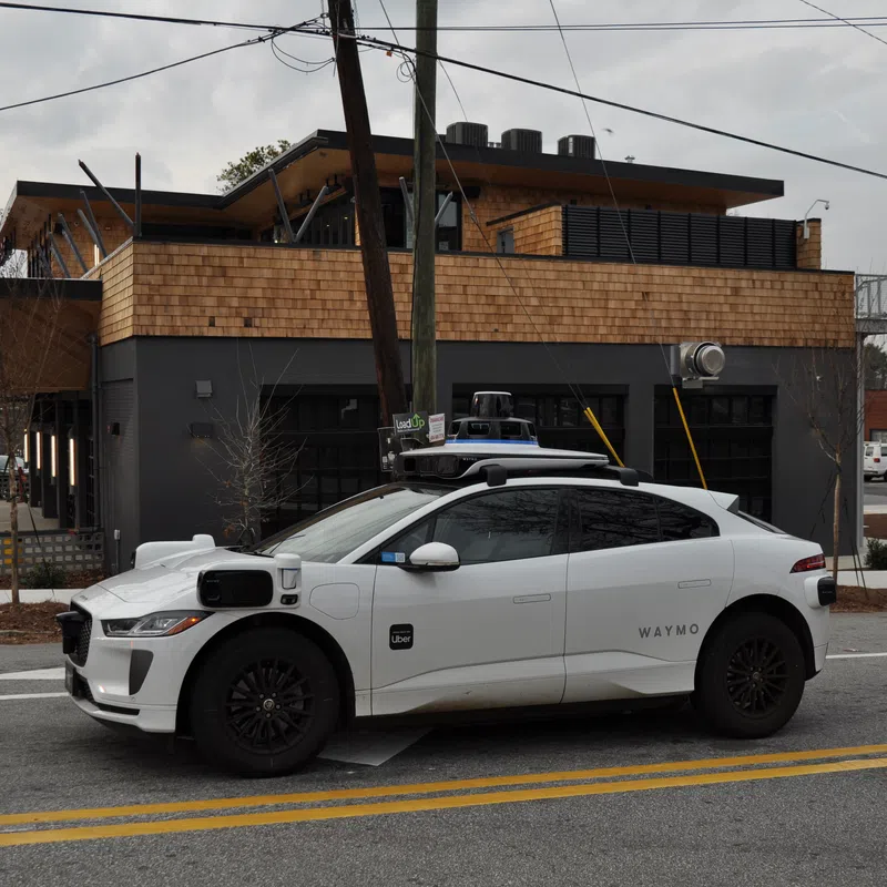 White Waymo self-driving electric car parked on a city street with modern building in the background.