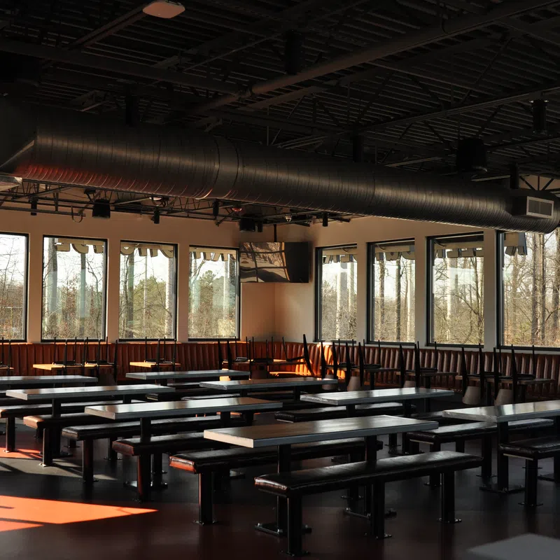 Empty modern cafeteria with large windows, wooden benches, and overhead ventilation ducts during daytime.