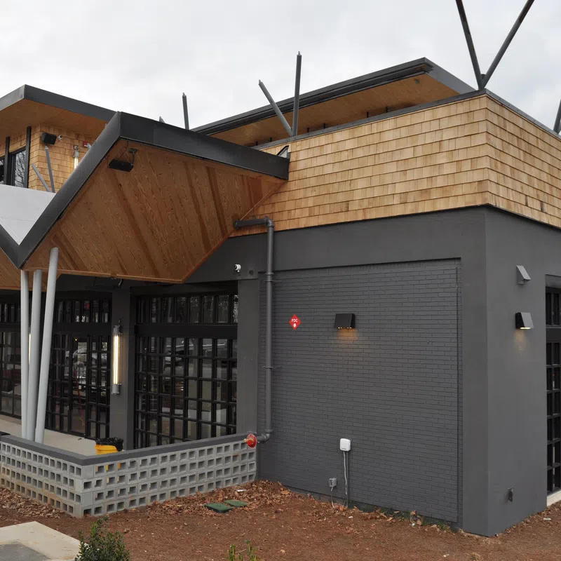Modern restaurant building with black brick walls, large windows, and wooden shingle roofing under gray sky