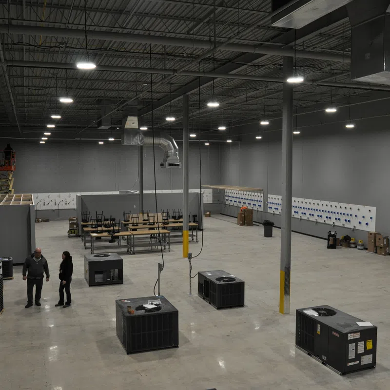 Large industrial warehouse with HVAC units, worktables, and two people standing on concrete floor under high ceiling lights.
