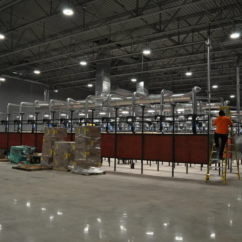 Large industrial warehouse with machinery, ventilation ducts, pallets, and workers performing maintenance tasks.