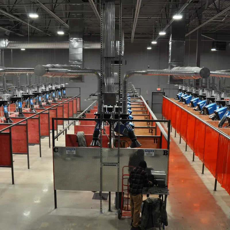Industrial welding station with multiple booths, blue welding machines, red curtains, and workers in a large factory.