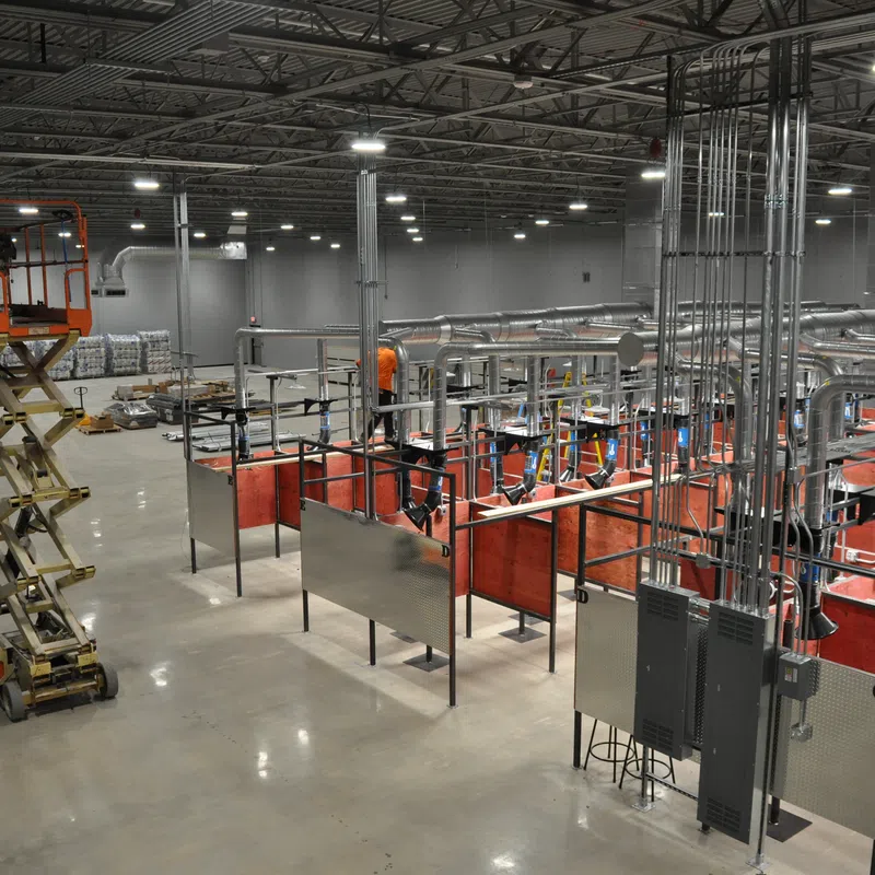 Worker on scissor lift inside large industrial workshop with welding stations and ventilation system.
