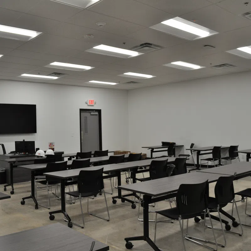 Spacious modern classroom with black chairs and tables, ceiling lights, TV screen, and white walls.