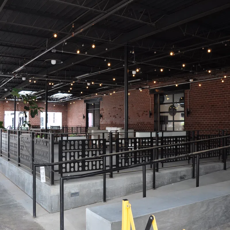 Modern indoor dining area with brick walls, metal railings, string lights, and concrete flooring in an industrial style.