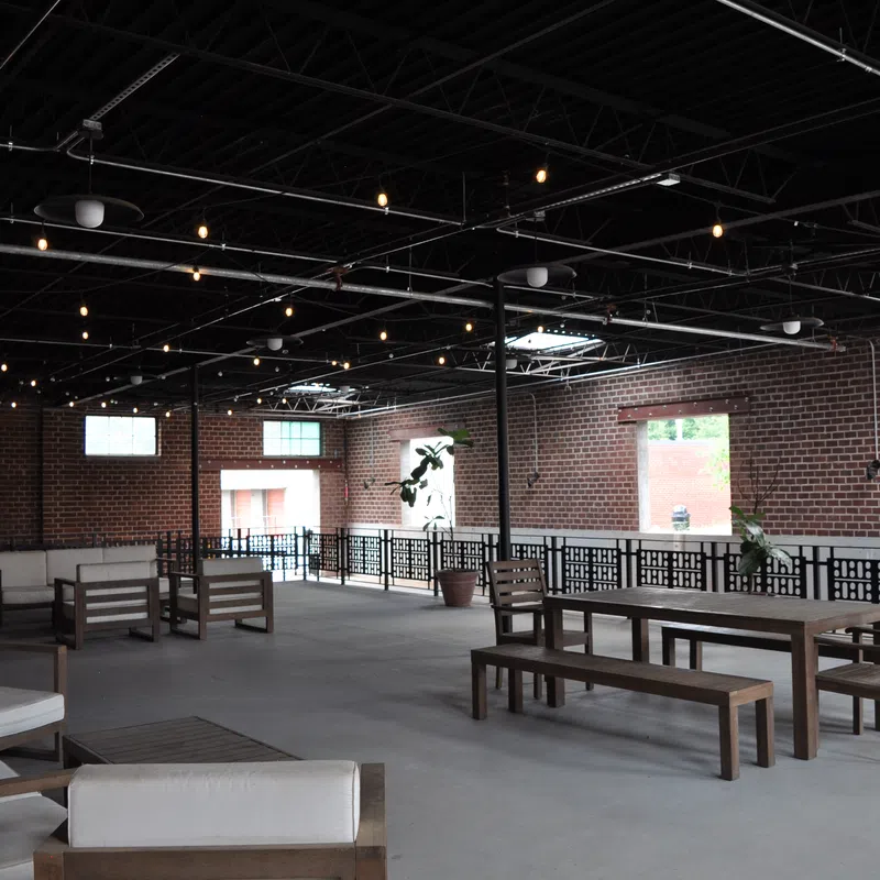Spacious indoor lounge with wooden furniture, exposed brick walls, and string lights under a high ceiling.