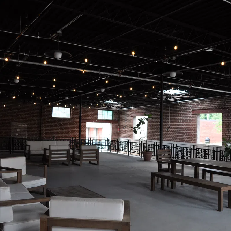 Spacious indoor patio with wooden sofas and tables, exposed brick walls, and string lights overhead.