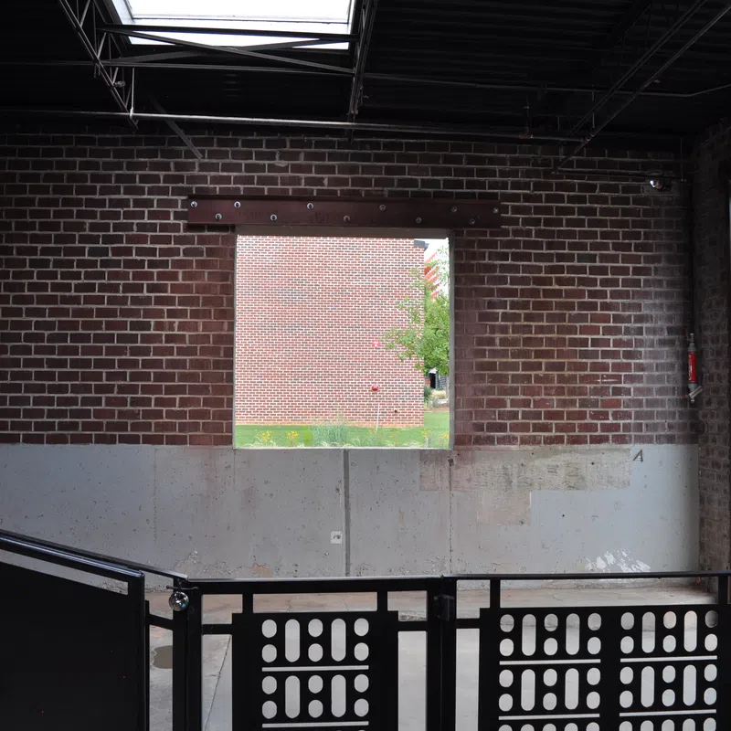 Interior view of an industrial-style building with brick walls, a square window, black railing, and concrete lower wall.
