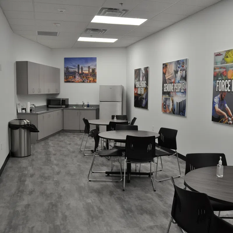 Modern office break room with round tables, black chairs, kitchenette, fridge, and motivational wall posters.