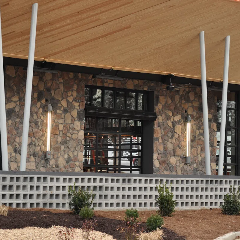 Modern building exterior with stone wall, large windows, vertical lights, and wooden roof overhang.