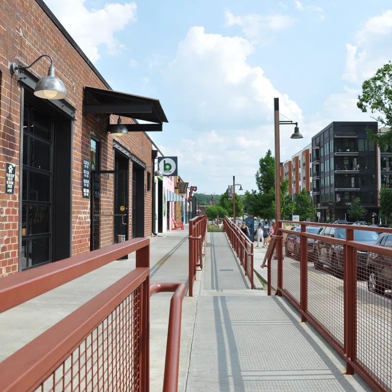 Street view of urban sidewalk with red railings, brick buildings, parked cars, and modern apartments under blue sky.