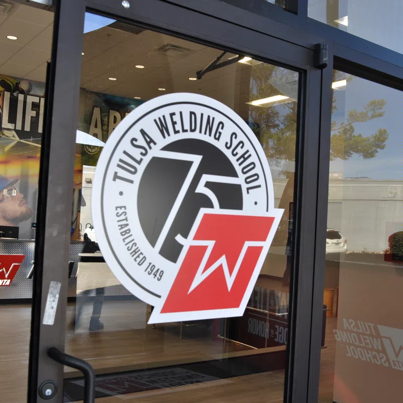 Glass door entrance of Tulsa Welding School with logo and view of interior training area with welding equipment.