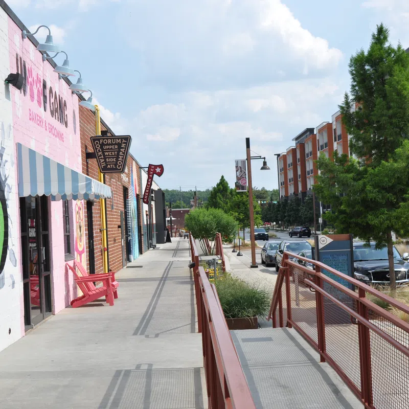 City sidewalk with colorful storefronts, trees, parked cars, and a clear sky on a sunny day