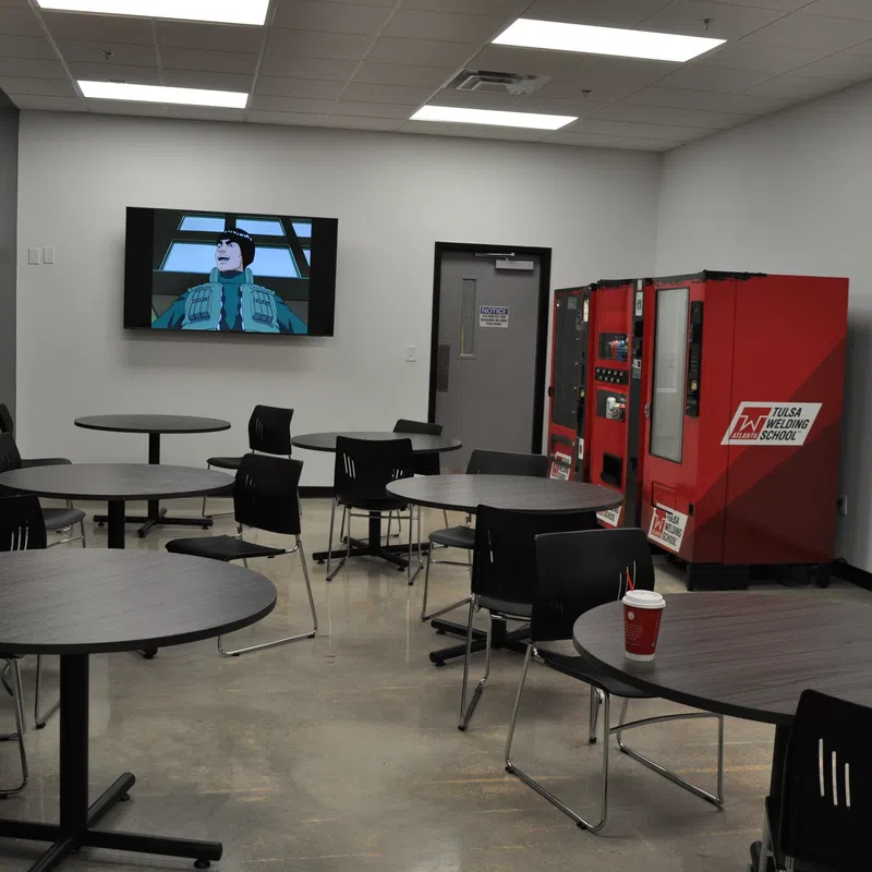 Modern break room with round tables, chairs, vending machines, hand sanitizer, and a TV mounted on the wall.
