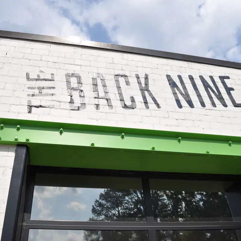 Exterior of a white brick building with The Back Nine sign and green metal awning under a partly cloudy sky