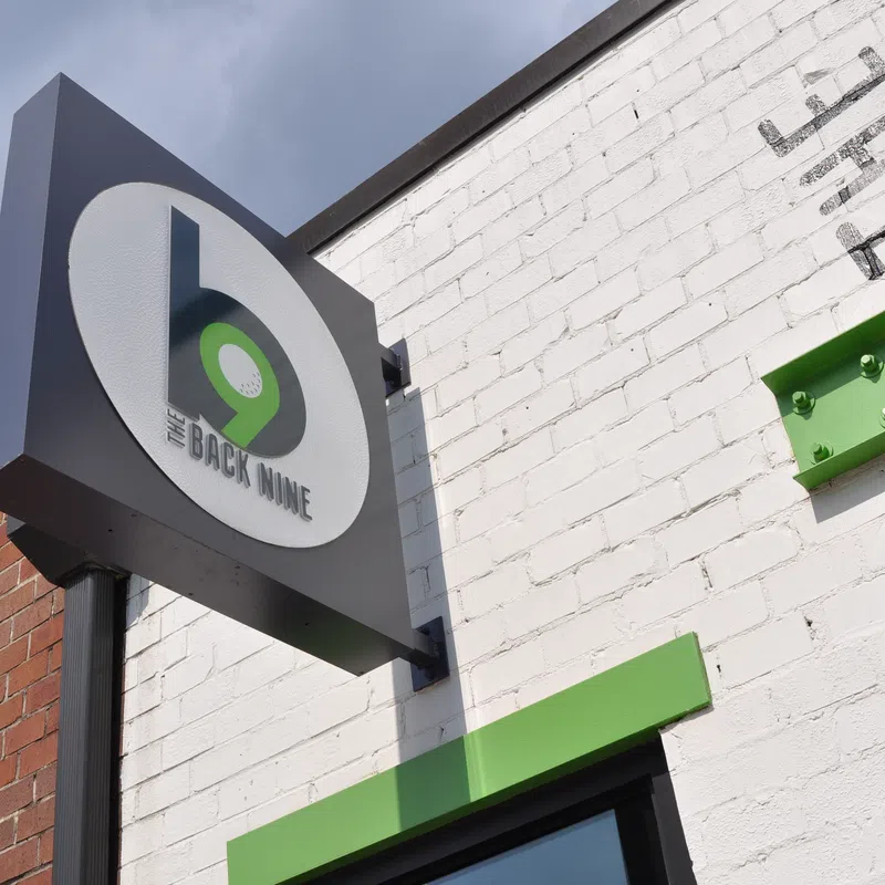 Exterior sign of The Back Nine bar with green and black logo on white and brick wall background.