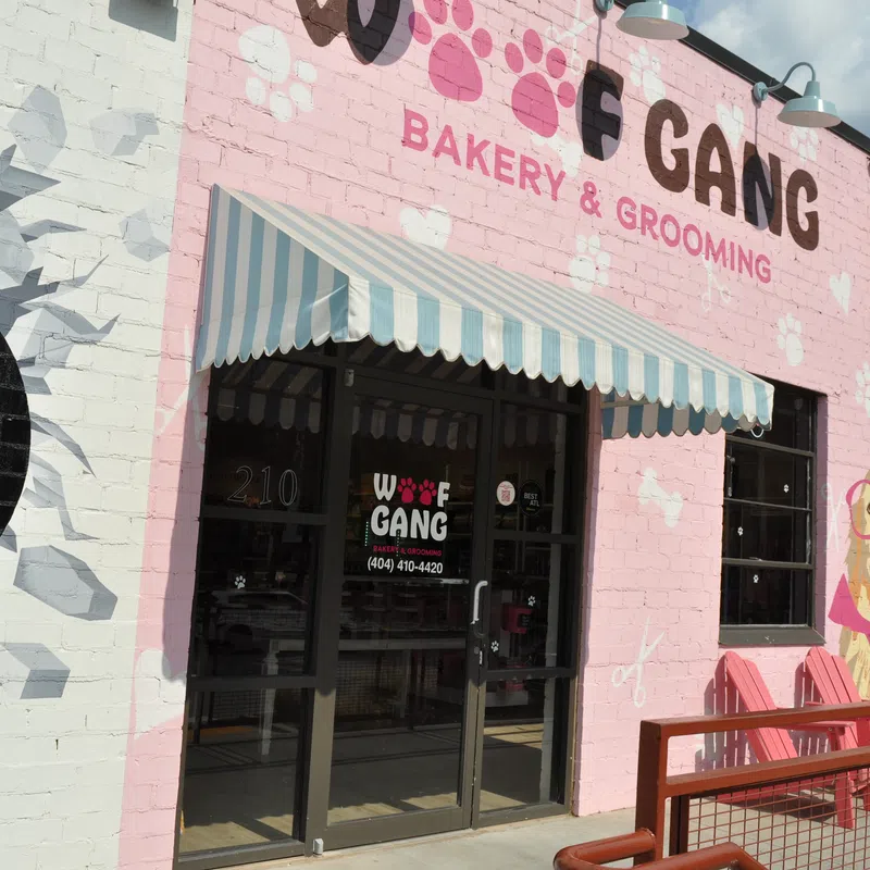 Entrance of Woof Gang Bakery & Grooming with pink mural, striped awning, and dog illustration on brick wall.