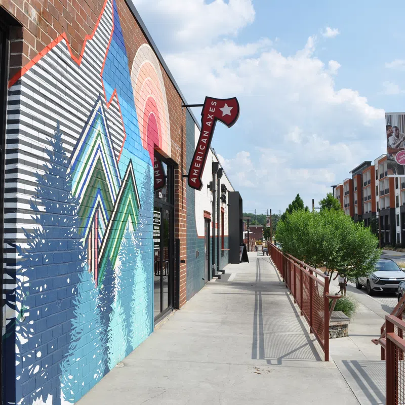 Street view of a colorful mural with geometric trees and rainbow over building, sidewalk, and modern apartments in background
