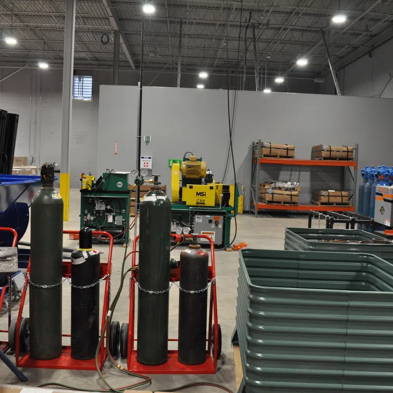 Industrial warehouse with gas cylinders, machinery, storage racks, and plastic containers under bright lighting.