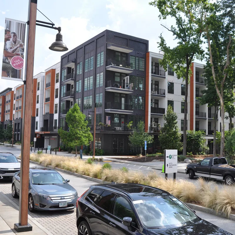 Modern apartment complex with parked cars, street lamps, trees, and landscaped sidewalks on a sunny day.